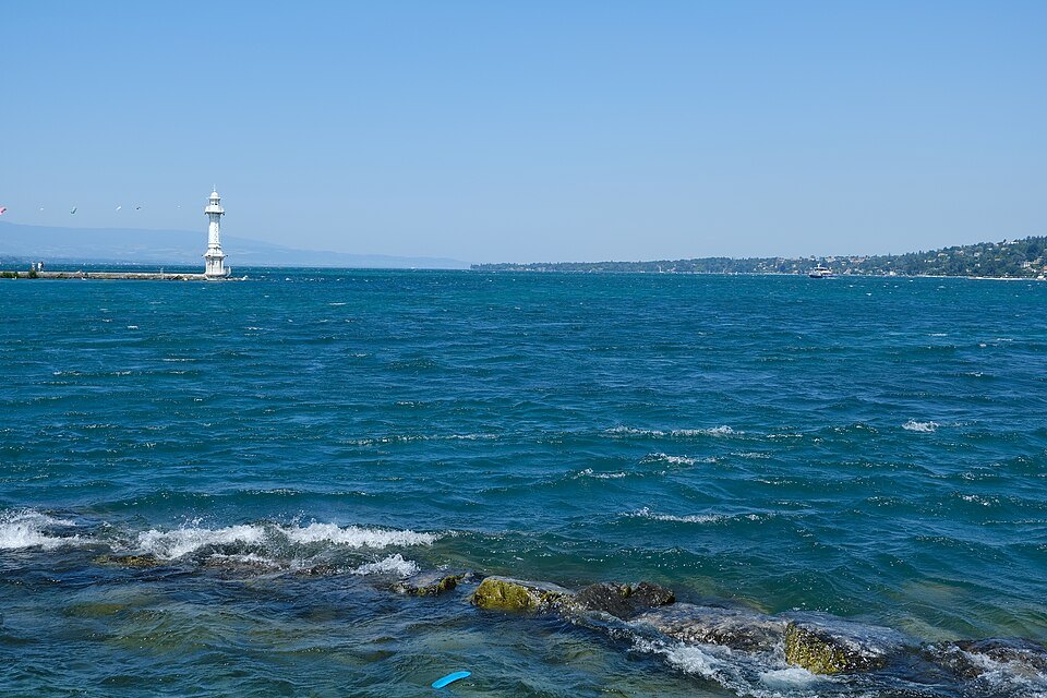 The white Pâquis lighthouse at the harbor entrance, waves in the foreground