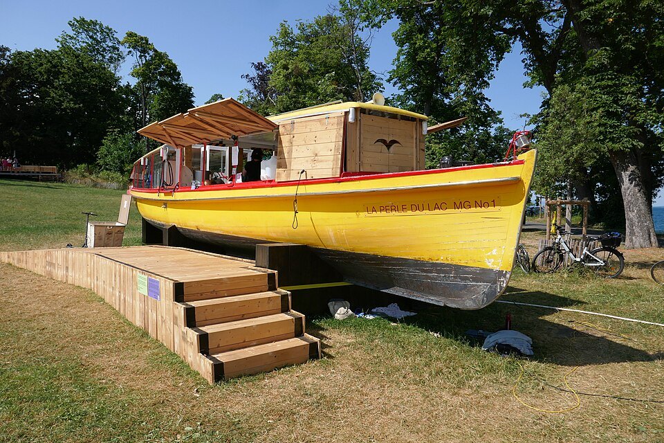A yellow wooden boat set on a platform on the lawn of the Perle du Lac