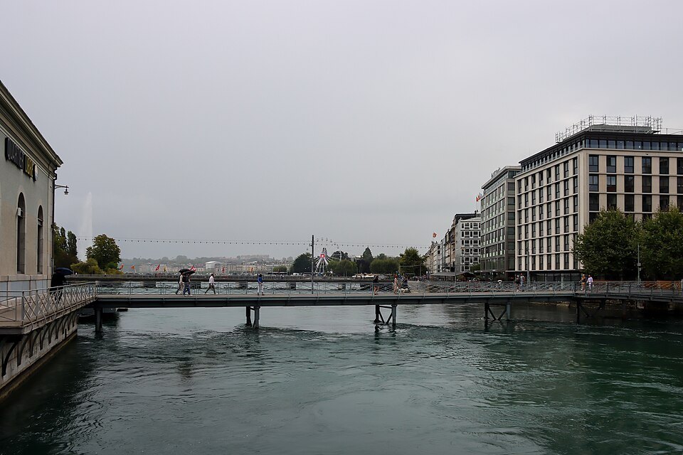 The Rhône and a footbridge under a gray sky, the Jet d'Eau faint in the distance