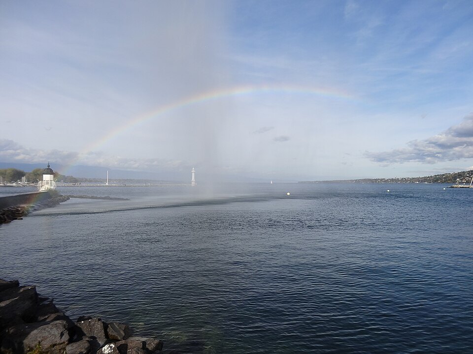 A faint rainbow in the spray of the Jet d'Eau over a gray harbor