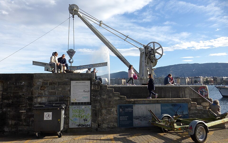An old harbor crane on a stone pier by the quay, people sitting beneath